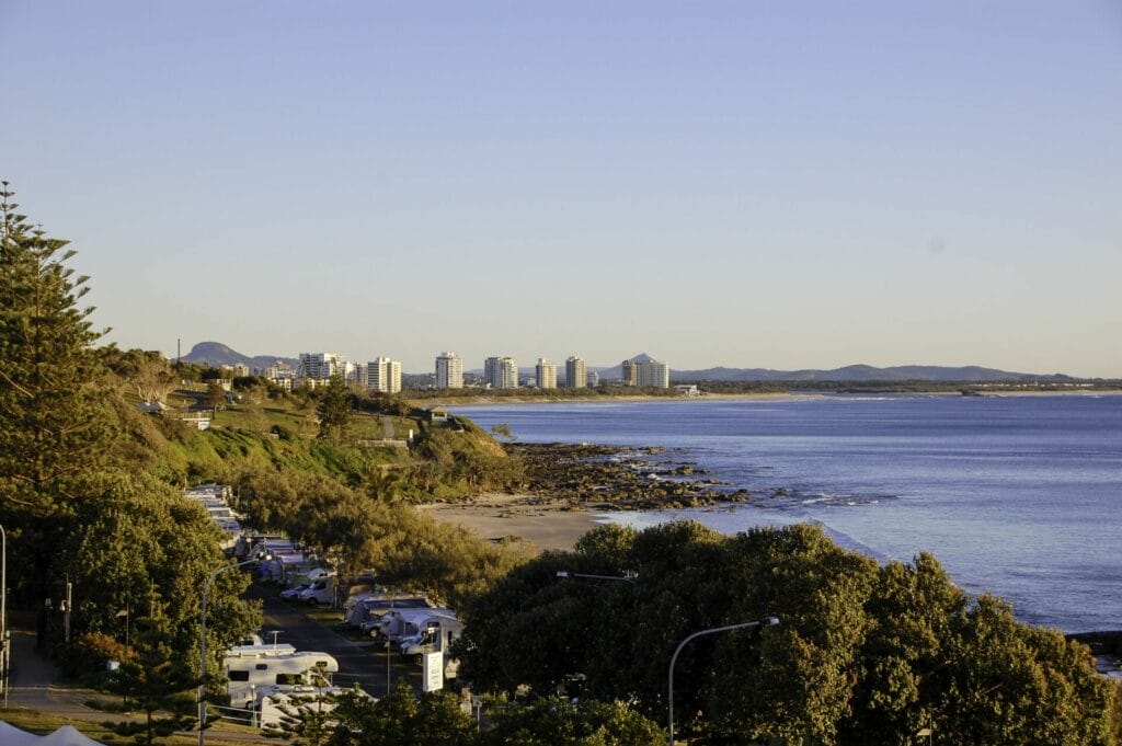 Inverness Coastal Cityscape With High-Rise Buildings, Rocky Shoreline, And Parked Cars Along A Tree-Lined Road At Sunset—Perfect For A Private City Tour Or Sightseeing Service.